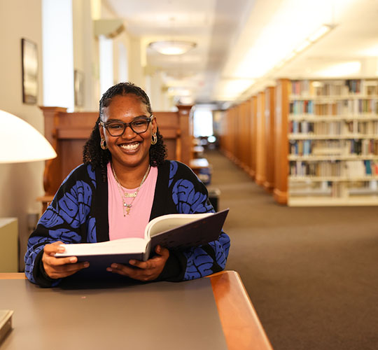 A student sitting in the Sawyer Library holding a book and smiling