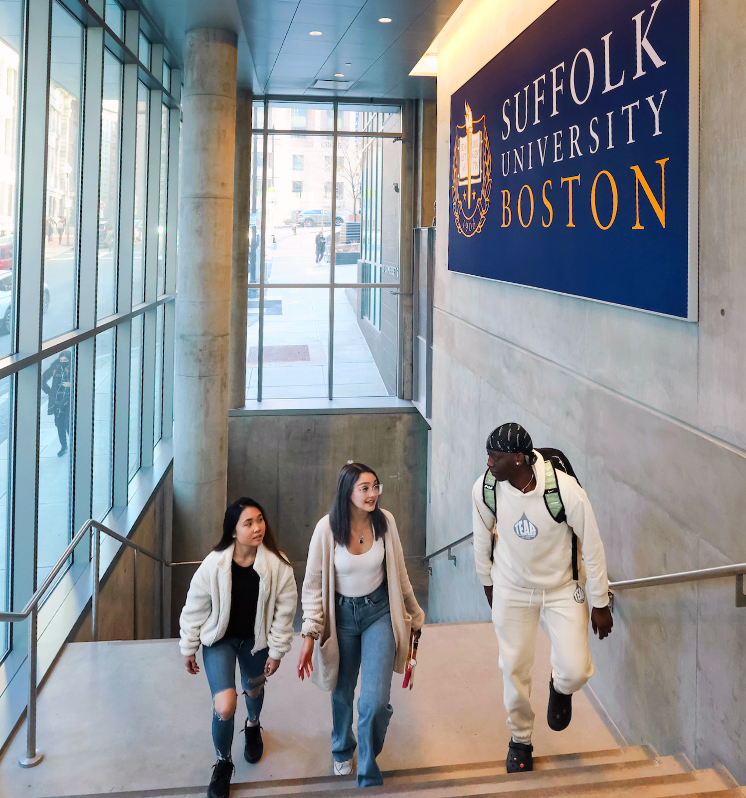 Three Suffolk students walk up the stairs in a campus building while talking together.