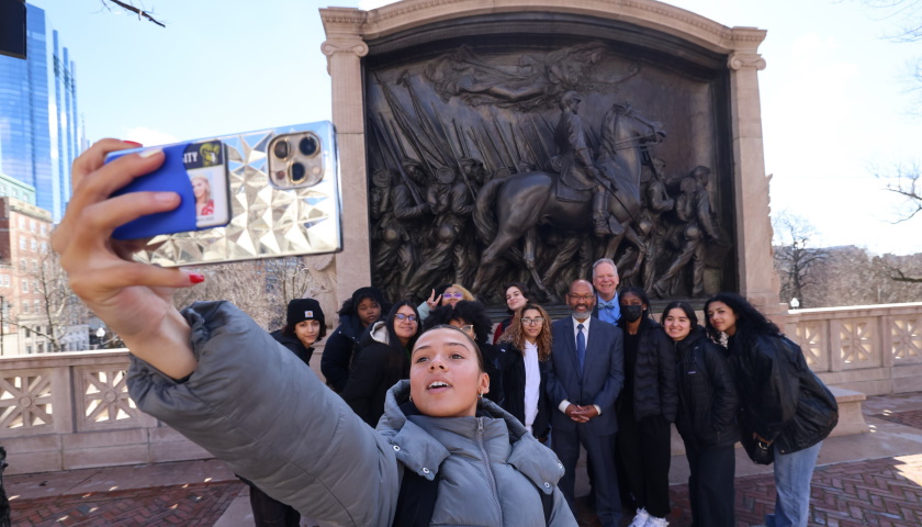 A student holds a phone to take a selfie with classmates and Professors Charles Yancey and Bob Allison in front of the Robert Gould Shaw and Massachusetts 54th Regiment Memorial