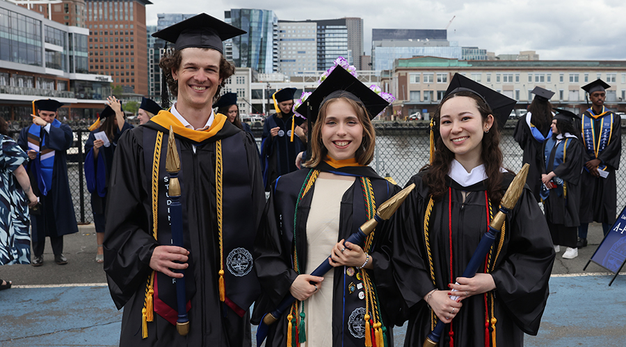 Three students in their caps and gowns holding the mace and other items ahead of leading the graduates of the College of Arts & Sciences to Commencement