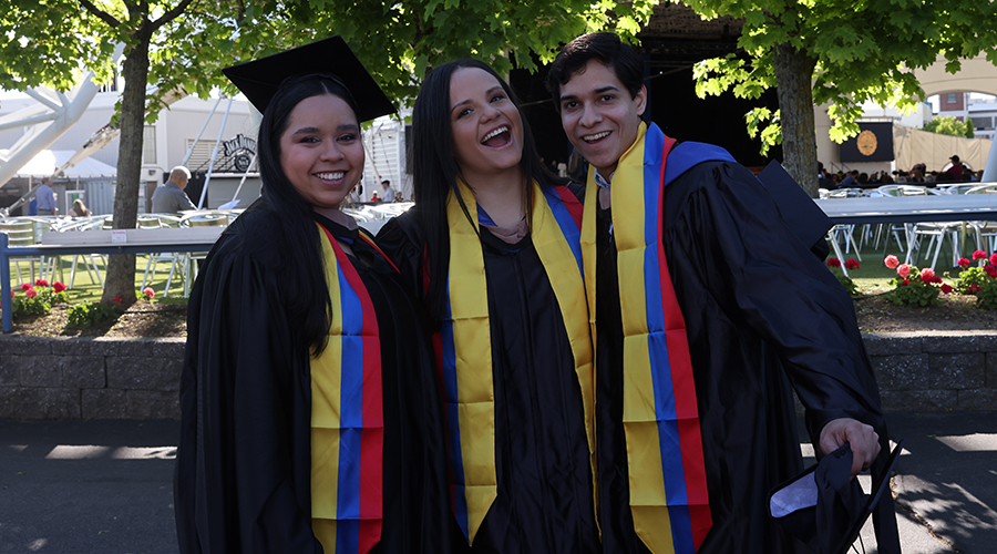 Three graduating students smile as they wait outside the Leader Bank Pavilion before Commencement 2025