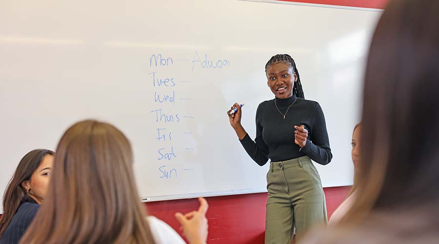 A student stands in front of a classroom whiteboard with a marker in hand, turning to look at her peers.