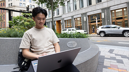 Suffolk student works on a laptop on a bench outside surrounded by Boston buildings.