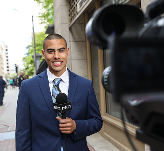 Student holds microphone as he reports from the street outside Studio 73