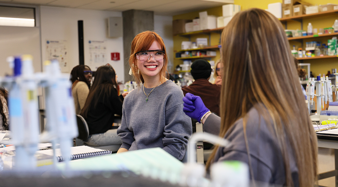 Two Suffolk students wearing gloves and goggles in discussion before class begins in a lab.