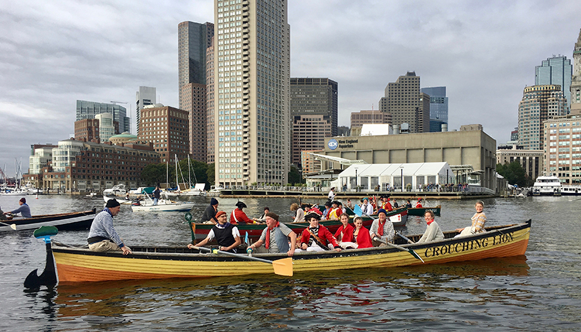 Redcoat reenactors paddle across Boston Harbor in a longboat, with modern skyscrapers providing a backdrop