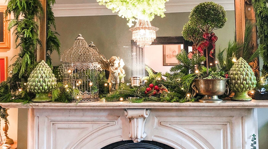 Mantelpiece decorated with greens, a bird cage, angel, topiary and more, all reflected in the mirror over the fireplace