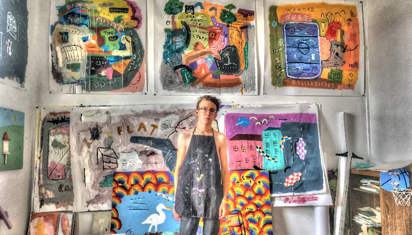 Cameron Boyce in black painter's apron with large, colorful artwork hanging on he wall behind him