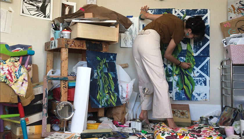 Feltoon painting green leaves over a blue-and-white background in her cluttered bedroom studio