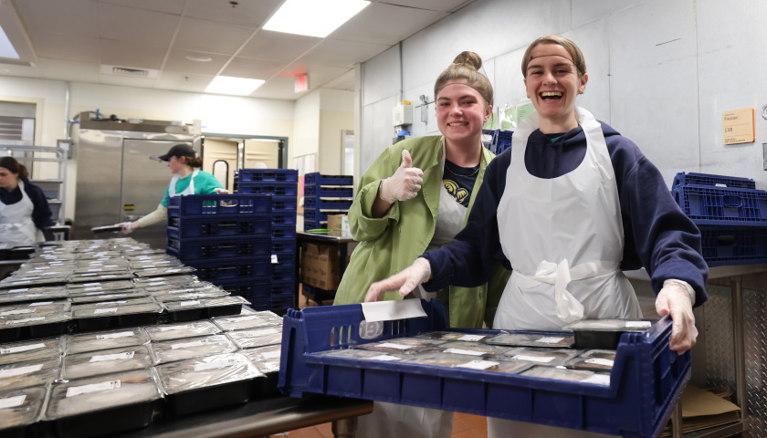 Two students in aprons work in the Community Servings kitchen; one carries trays of prepared food while the other gives a thumbs up