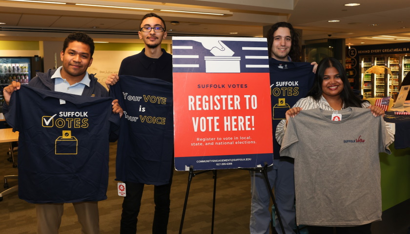 Four students hold us Suffolk Votes t-shirts while standing next to a voter registration sign
