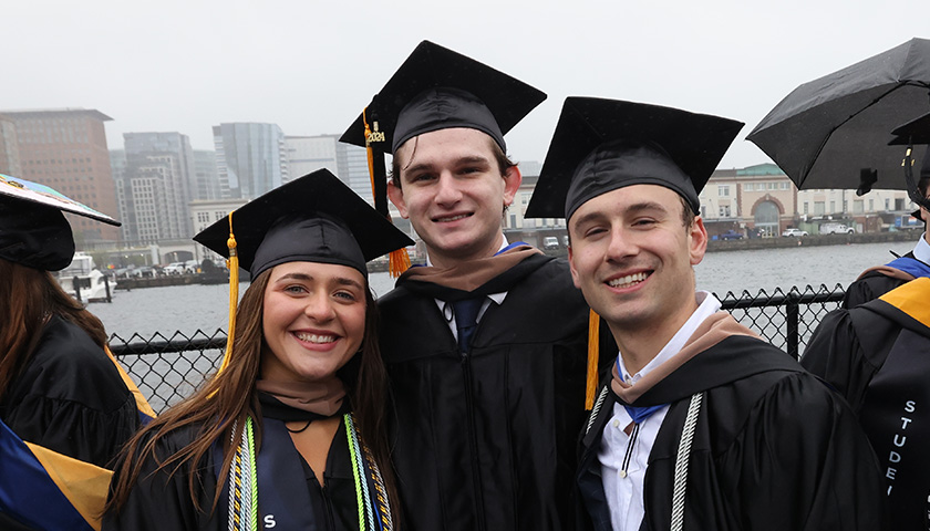 Three students pose in their caps and gowns outside the Leader Bank Pavilion by the water
