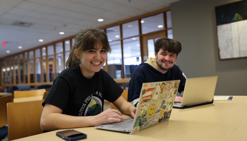 Juliette Salah and Will Woodring smile as they sit with their laptops open in the library