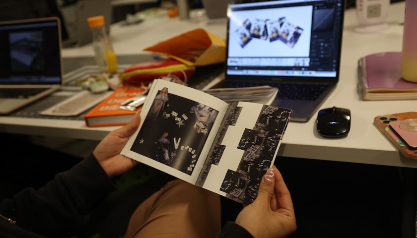 Hands hold open a physical booklet as the same artwork appears on a laptop screen beyond