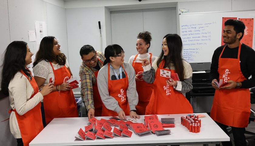 Students in the Publication Design course wearing the group's orange aprons, smiling and laughing together in the design lab