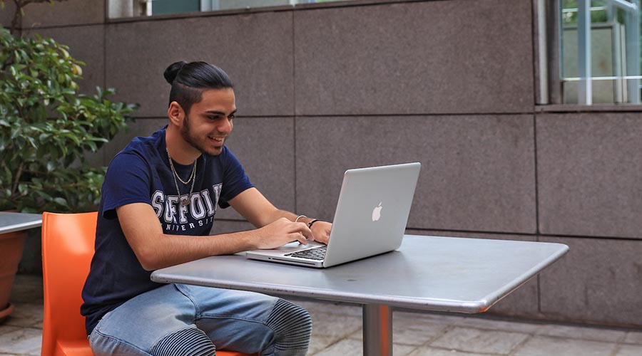 A Suffolk student sits at an outdoor table with his laptop.