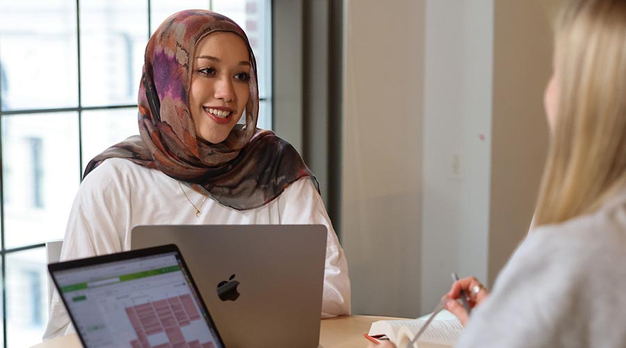 A Suffolk student sits smiling in a meeting in front of windows with laptops on the table.